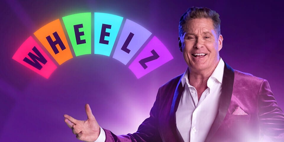 Programvert som gestikulerer mot et fargerikt hjul med teksten «Wheelz Casino» over, satt mot et lilla bakteppe.