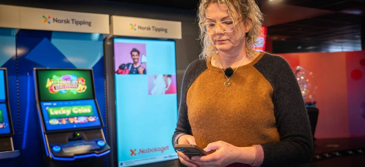 En person med briller og krøllete hår står innendørs og bruker en telefon, med Norsk Tipping-spillautomater i bakgrunnen, og reflekterer over den pågående norsk tipping-tillittskrisen.